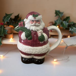 May include: A ceramic Santa Claus mug with a white handle. The mug is in the shape of Santa, wearing a red suit with white trim, a matching hat, and black boots. He holds a green sack of gifts and a candy cane.