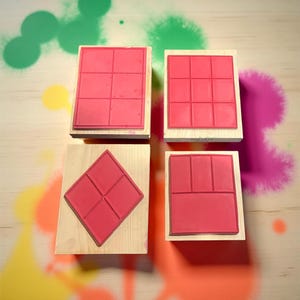 May include: Four red rubber stamps with various grid patterns mounted on wooden blocks. The stamps are arranged on a light-colored surface with colorful paint splatters in the background. The stamps are for crafting or scrapbooking.