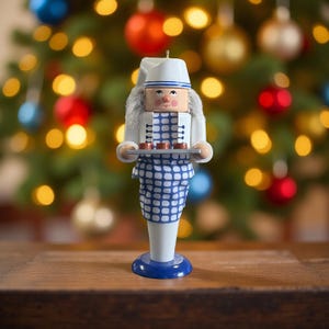 Op de afbeelding: Een houten kerstornament in de vorm van een kok, met een witte hoed en jas en een blauw-wit geruite schort. De kok houdt een dienblad met kleine, bruine lekkernijen vast. Het ornament heeft een blauwe basis en staat tegen een vage kerstboomachtergrond.