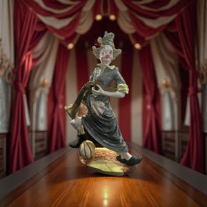 Peut inclure: Figurine de clown en céramique dans un cadre de cirque. Le clown porte une tenue grise et noire, tenant un instrument de musique. La figurine est posée sur une surface en bois, avec un fond de rideaux rayés rouges et blancs.
