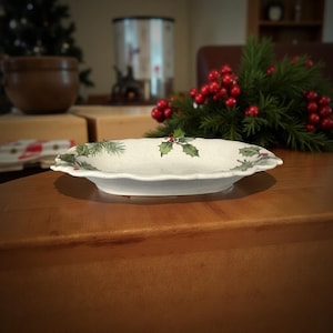 Puede incluir: Un plato ovalado de cerámica blanca con borde ondulado. El plato está decorado con ramitas verdes y bayas rojas, evocando un tema festivo navideño. El plato está colocado sobre una superficie de madera.