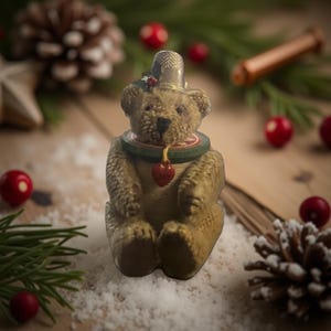 Puede incluir: Pequeña figura de oso de peluche marrón claro con un collar verde y rojo y un sombrero plateado. Un colgante de corazón rojo cuelga del collar. El oso está rodeado de decoraciones navideñas, incluyendo piñas y bayas rojas.