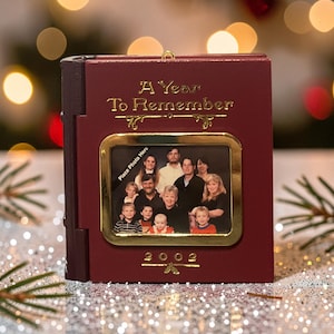 Puede incluir: Un álbum de fotos de color burdeos con un marco dorado y el texto "A Year To Remember" y "2002". El álbum contiene una foto familiar. También son visibles las palabras "Place Photo Here".