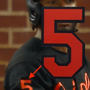 May include: A black baseball jersey with orange trim and the number "5" on the sleeve and back. The jersey has the word "Orioles" written in orange on the front.