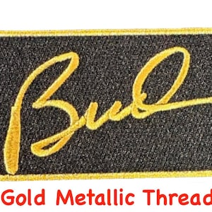 May include: A black rectangular patch with a gold embroidered signature that reads "Bud" in a cursive font. The patch has a gold metallic thread border.