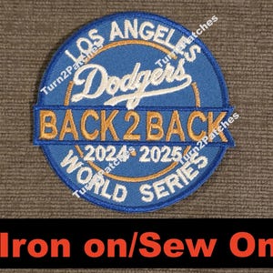 May include: A blue and white embroidered patch featuring the text "Dodgers" and "BACK 2 BACK" in gold. The patch also includes the text "LOS ANGELES" and "WORLD SERIES" in white. The bottom of the patch reads "Iron on/Sew On".