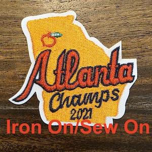 May include: An iron-on or sew-on patch featuring an orange outline of the state of Georgia with the word "Atlanta" in red and black lettering and the words "Champs 2021" in black lettering.
