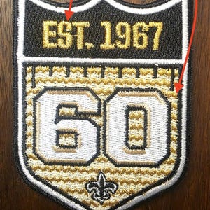 May include: A shield-shaped embroidered patch featuring the text EST. 1967 in gold metallic thread at the top. The center displays the number 60 in white with black outlines, set against a gold textured background with a fleur-de-lis symbol below.