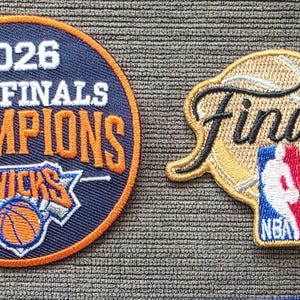 May include: A circular blue patch with an orange border featuring the text 2026 NBA FINALS CHAMPIONS KNICKS and a basketball graphic, alongside a gold-toned patch shaped like a basketball with the word Finals and the NBA logo in red, white, and blue.