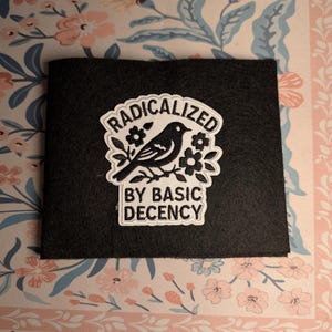 May include: A black felt patch with the text "RADICALIZED BY BASIC DECENCY" in a white, stylized font. The patch features a black bird perched on a branch with white flowers. The background is a floral pattern.