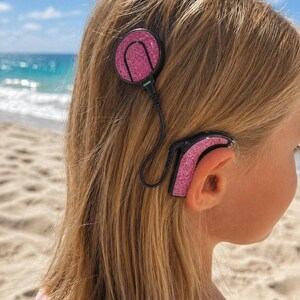 Könnte beinhalten: Ein rosa glitzerndes Cochlea-Implantat ist am Kopf einer Person befestigt. Das Gerät hat eine kreisförmige Komponente und eine am Ohr getragene Komponente, die durch ein schwarzes Kabel verbunden sind. Der Hintergrund zeigt einen Strand mit Sand und Meer.