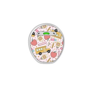 May include: A round, silver-edged device with a pink background featuring a repeating pattern of school-themed illustrations. The pattern includes a yellow school bus, red apples, pencils, glue bottles, and rulers. A green light is visible on the top of the device.