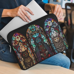 May include: A black laptop sleeve with a stained glass design featuring three cats. The cats are depicted in a gothic style with colorful stained glass windows behind them.
