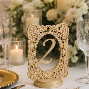 May include: A gold-colored table number sign featuring the numeral 2 centered within an oval frame. The frame is decorated with intricate, raised scrollwork and floral-inspired relief patterns, supported by a solid, rounded base.