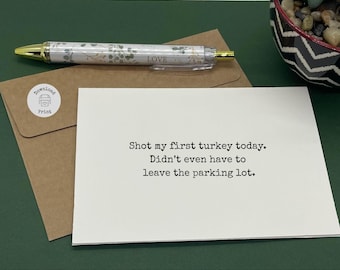 May include: A humorous greeting card with the text "Shot my first turkey today. Didn't even have to leave the parking lot." is next to a pen with a floral design and the word "LOVE". A brown envelope and a small bowl of stones are also in the image.