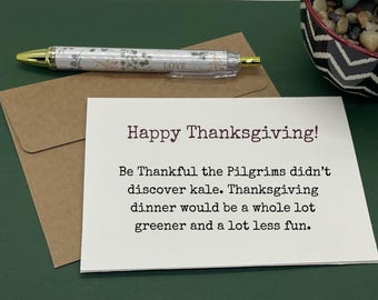 May include: A Thanksgiving card with the message "Happy Thanksgiving!" and a humorous text about kale. A pen with a floral pattern and a gold tip lies on a brown envelope. The background is a dark green surface.