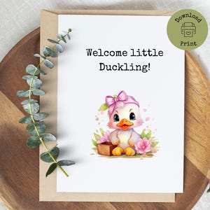Könnte beinhalten: Eine Grußkarte mit dem Text "Welcome little Duckling!". Sie zeigt ein Cartoon-Entchen mit rosa Schleife und rosa Blumen. Die Karte liegt auf einem Holztablett mit Eukalyptus.