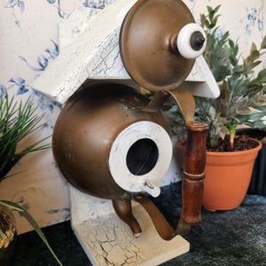 Könnte beinhalten: Ein Vogelhaus aus einer wiederverwendeten Kupferkanne mit einem weiß gestrichenen Dach und einem Holzgriff. Das Vogelhaus hat ein rundes Loch, durch das die Vögel eintreten können.