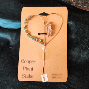 May include: Copper plant stake featuring a heart-shaped design. The copper wire heart is decorated with turquoise beads and a quartz crystal pendant. The stake is attached to a brown cardboard backing with the words "Copper Plant Stake". A small tag says "remove".