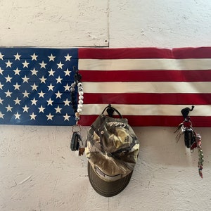 May include: A wooden American flag painted in red, white, and blue with three hooks hanging from the bottom. A camouflage baseball cap hangs from one hook, and a set of keys hangs from another hook.