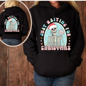 May include: Black hoodie with a graphic of a skeleton wearing a Santa hat and holding a disco ball. The text "Just Waiting for Christmas" is written in a retro font around the skeleton.