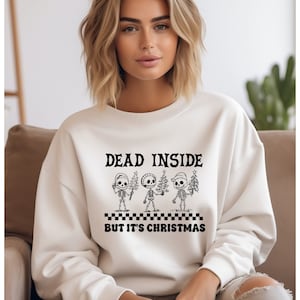 May include: White sweatshirt with a black graphic print of three skeletons holding Christmas trees. The text "Dead Inside But It's Christmas" is printed below the skeletons.