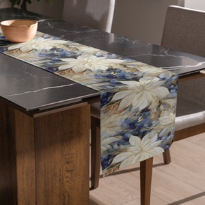 May include: A blue and beige floral table runner with a modern design. The runner is placed on a black table with a wooden base.