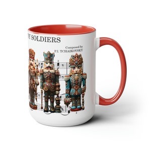 May include: White ceramic mug with a red rim and handle. The mug features a design of four nutcracker soldiers in front of sheet music with the text "Toy Soldiers" and "Composed by P.I. Tchaikovsky".