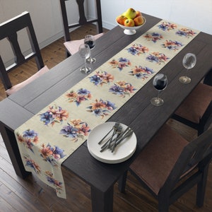 Può includere: Un runner da tavolo beige con motivi floreali ad acquerello nei toni del blu, viola e pesca. Il runner è posizionato su un tavolo di legno scuro apparecchiato per un pasto, con piatti, posate e calici.