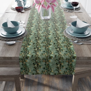 May include: A green table runner featuring a repeating floral vine pattern. The runner is laid on a wooden table, set with plates, bowls, cutlery, and wine glasses. A vase of pink lilies is positioned in the centre of the table.