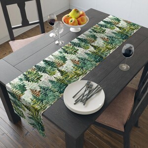 May include: A dark wood dining table set with a forest-themed table runner. The runner features a repeating pattern of green and white pine trees. The table is set with white plates, silverware, wine glasses, and a bowl of apples.