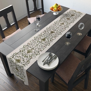 May include: A cream-colored table runner with a detailed green and gold floral design, placed on a dark wooden table. The table is set with wine glasses, plates, and silverware, and a bowl of fruit.