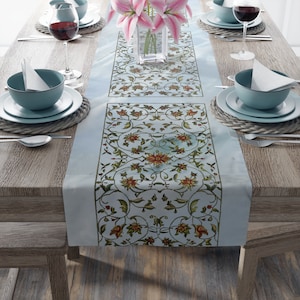 May include: A light blue table runner with a floral design in red, green, and gold. The runner is placed on a wooden table set with dishes, silverware, and wine glasses. A vase of pink flowers sits in the center.
