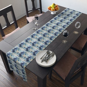 May include: A rectangular table runner with a blue, white, and gold abstract pattern. The runner is placed on a dark wooden table set for a meal, with plates, silverware, and wine glasses. A bowl of fruit sits at the table's center.
