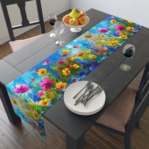Può includere: Un runner da tavolo floreale colorato con sfondo blu e fiori gialli, rosa e viola. Il runner è su un tavolo in legno scuro con due bicchieri di vino e una ciotola di mele.