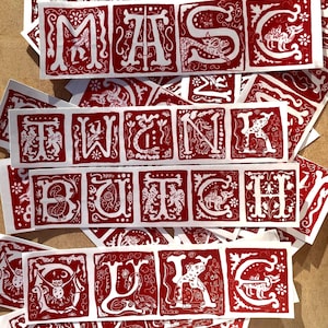 Puede incluir: Pegatinas rojas y blancas con las palabras "MASK", "WANK", "BUTCH" y "LUKE" en una fuente decorativa de estilo medieval. Cada letra está dentro de un cuadrado rojo, con ilustraciones blancas. Las pegatinas están esparcidas sobre una superficie de madera.