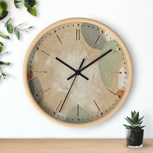Pode incluir: Um relógio de parede redondo com moldura de madeira clara. O mostrador do relógio tem um fundo texturizado e neutro com formas abstratas em cores suaves. Os ponteiros pretos indicam as horas. O relógio tem aproximadamente 30 cm de diâmetro.