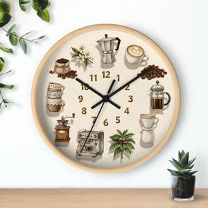 Op de afbeelding: Ronde wandklok met een licht houten frame en een crèmekleurige wijzerplaat. De wijzerplaat toont illustraties van koffiegerelateerde items, waaronder een koffiezetapparaat, bonen en kopjes. Zwarte wijzers geven de tijd aan.