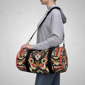 May include: A black duffel bag with a colorful print featuring a cat's face, floral patterns, and an eye. The bag has a black strap and a zipper closure.