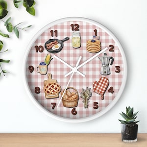 Puede incluir: Un reloj de pared redondo con marco blanco y fondo de cuadros vichy rosa y blanco. La esfera presenta ilustraciones de desayunos, una cafetera, una cesta de pan y un ramo de flores, con números de color marrón oscuro del 1 al 12.