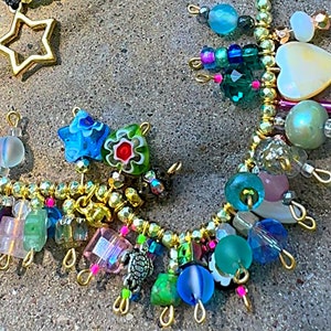 Puede incluir: Una colección de collares de cuentas de colores. Un collar presenta un colgante de estrella dorada. Otros collares tienen una variedad de cuentas en tonos azules, verdes, rosas y transparentes, con detalles dorados. El texto "Rock Candy Bar" es visible.