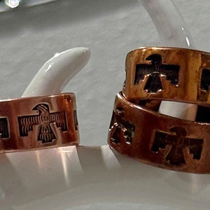 Pode incluir: Dois anéis de cobre com um design de pássaros estilizados. Os anéis estão empilhados um em cima do outro.