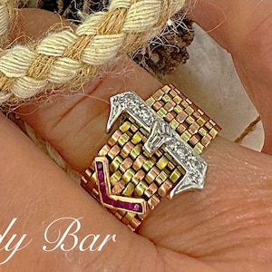 May include: A gold ring with a braided design featuring a red stone and a diamond. The ring is on a finger against a background of a pine branch and a pine cone.