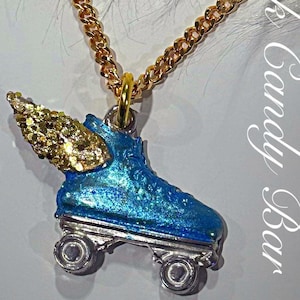 Puede incluir: Un collar de cadena dorada con un dije de patín de ruedas azul con un ala de brillantina dorada. El dije tiene ruedas plateadas. El texto "Rock Candy Bar" es visible en el fondo.