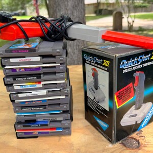 May include: A stack of ten grey Nintendo NES game cartridges including Excitebike, Monopoly, Wheel of Fortune, The Blue Marlin, Days of Thunder, Spy Hunter, Ghosts 'n Goblins, Captain Skyhawk, Gremlins 2, and a light gun controller with a Quick Shot XII box.