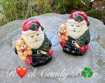 Könnte beinhalten: Zwei Keramikfiguren des Weihnachtsmanns, jede mit einem Teddybären. Die Weihnachtsmänner tragen rote Hüte mit Stechpalmen, grüne Mäntel, schwarze Stiefel und haben weiße Bärte. Die Teddybären sind hellbraun mit roten Fliegen. Die Figuren stehen auf einer grauen Steinoberfläche.
