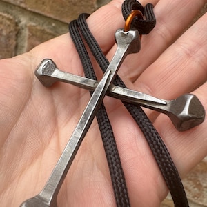 May include: A metal cross pendant made from nails, with a black cord. The cross has a rustic, industrial appearance, with a textured surface. The cord is knotted at the top and attached to a small orange ring.