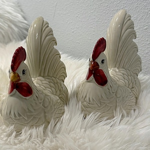 May include: Two white ceramic rooster figurines with red and yellow accents. The roosters are standing on a white fluffy surface.