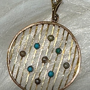 K&ouml;nnte beinhalten: Ein goldfarbener Anh&auml;nger mit einem kreisf&ouml;rmigen, gestreiften Design. Der Anh&auml;nger hat kleine, runde t&uuml;rkisfarbene und graue Steine. Eine kleine Perle ist in der dekorativen &Ouml;se oben eingesetzt. Der Anh&auml;nger ist wahrscheinlich Vintage oder antik.