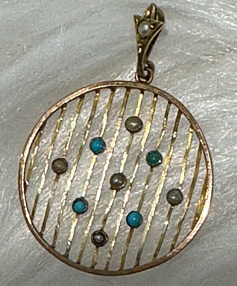 K&ouml;nnte beinhalten: Ein goldfarbener Anh&auml;nger mit einem kreisf&ouml;rmigen, gestreiften Design. Der Anh&auml;nger hat kleine, runde t&uuml;rkisfarbene und graue Steine. Eine kleine Perle ist in der dekorativen &Ouml;se oben eingesetzt. Der Anh&auml;nger ist wahrscheinlich Vintage oder antik.